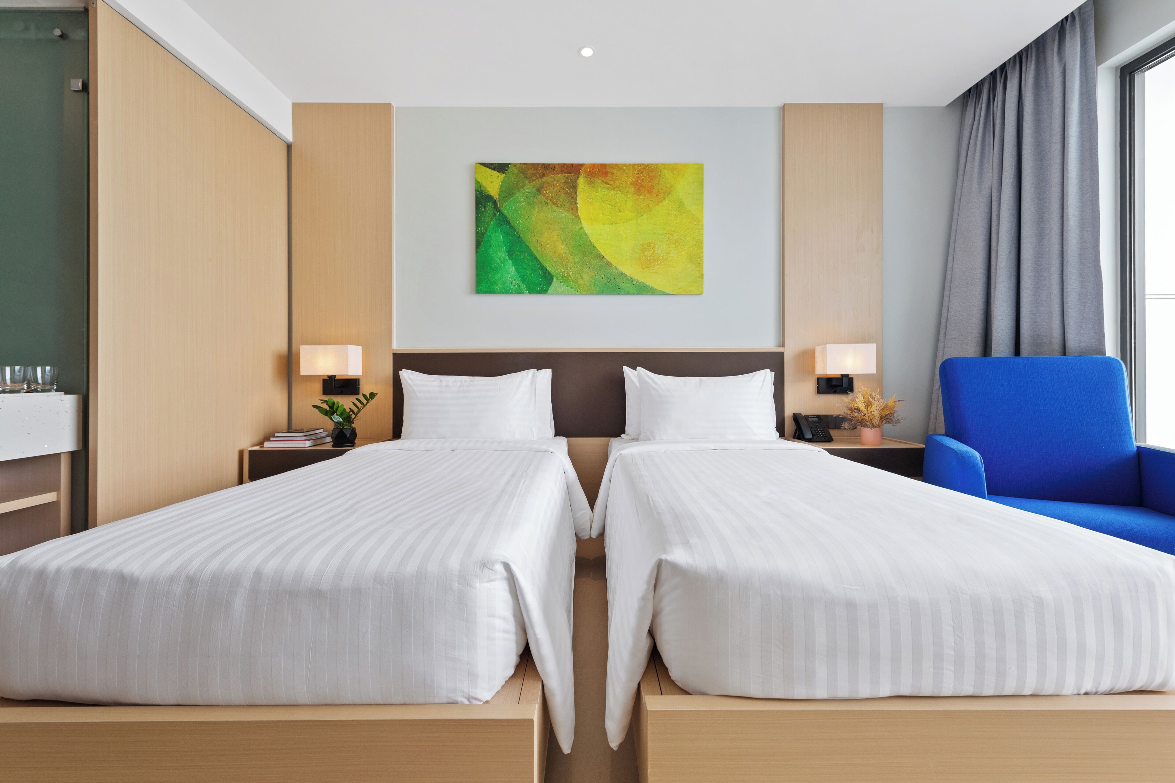 Une chambre d’hôtel avec deux lits, une grande peinture abstraite au mur, un fauteuil bleu et une vue sur l’extérieur.