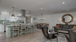A modern kitchen with a central island, dining area with wooden chairs, a grey sofa, and a round mirror on the wall.