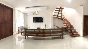 Amplia sala de estar con escalera de madera, un televisor de pantalla plana grande montado en la pared y un conjunto de sofás.