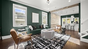 Sala moderna com parede verde, piso de madeira, sofá verde, poltrona de couro marrom, mesa de centro redonda e área de cozinha visível através de uma porta aberta.