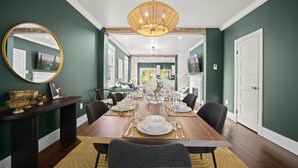 Uma sala de jantar com uma longa mesa de madeira posta para seis pessoas, rodeada por cadeiras estofadas escuras.