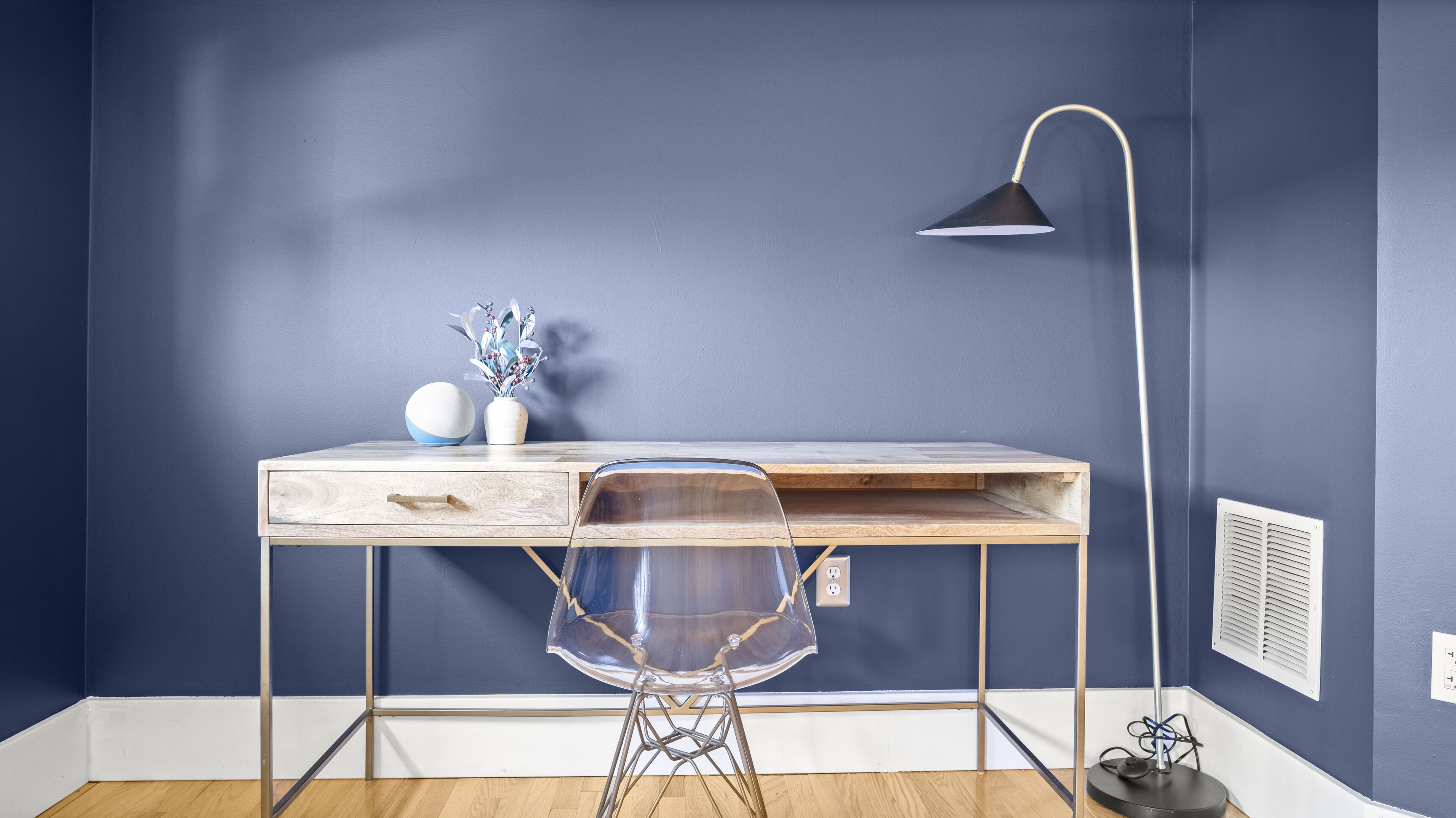 Un espace de travail moderne avec un bureau en bois, une chaise en plastique transparent, une lampe de bureau noire et un vase avec des fleurs.