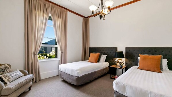 A hotel room with two beds, a sofa, a window with curtains, a chandelier, and a nightstand with a lamp.