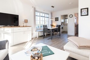 Ein modernes Wohnzimmer mit einer Couch, einem Esstisch mit Stühlen, einer Küchenzeile mit Fenster und einem Fernseher auf einem Schrank.