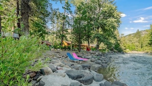 A riverside area with a rocky shore, a blue and pink lounge chair, and a wooden cabin in the background.