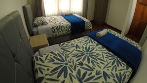 Quarto de hotel com duas camas, cada uma com colcha floral azul e branca, um guarda-roupa de madeira e uma janela com cortinas leves.