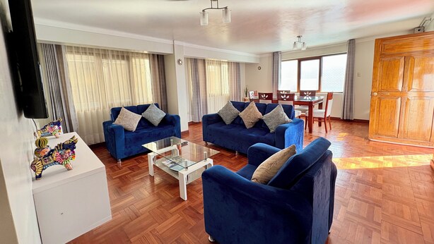 Sala de estar com dois sofás azuis, uma mesa de centro de vidro e uma mesa de jantar de madeira com cadeiras.