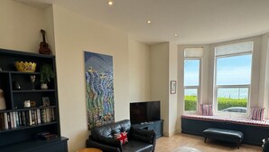 A living room with a bookshelf, a black leather sofa, a television, and a large window with a view of greenery. - Eva's Sea View 2 Bed 2 Bath Parking & NO GUEST FEES (Broadstairs)