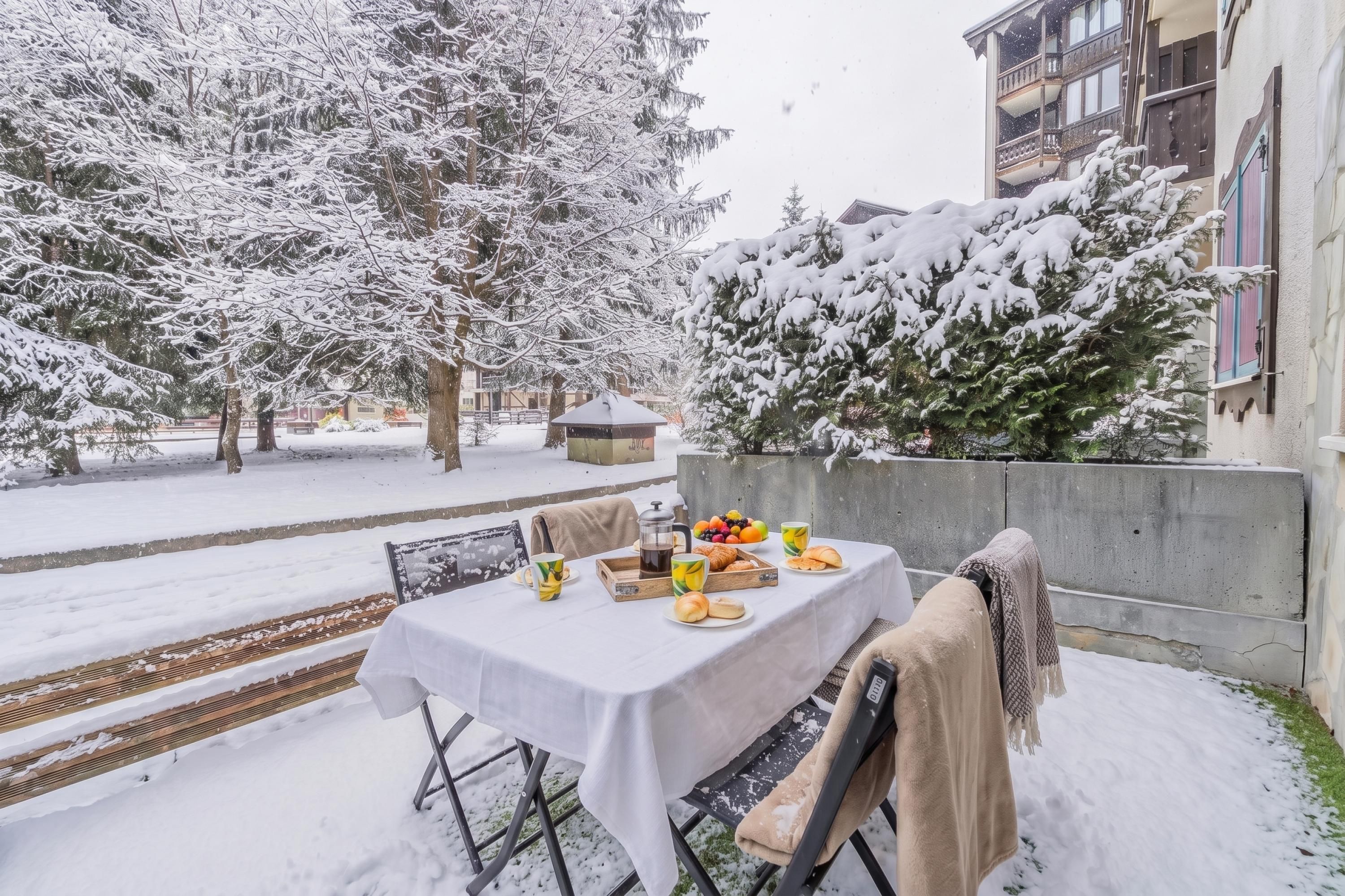 Un espace extérieur aménagé pour les repas, recouvert de neige, avec une table dressée pour deux personnes, entouré d’arbres chargés de neige et d’un bâtiment.