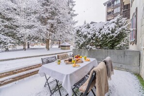 Un espace extérieur aménagé pour les repas, recouvert de neige, avec une table dressée pour deux personnes, entouré d’arbres chargés de neige et d’un bâtiment.