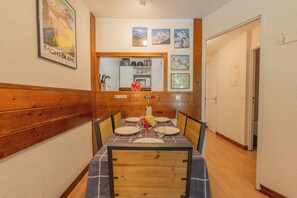 Un coin repas avec une table en bois dressée pour quatre personnes, une cuisine en arrière-plan et des tableaux encadrés accrochés au mur.