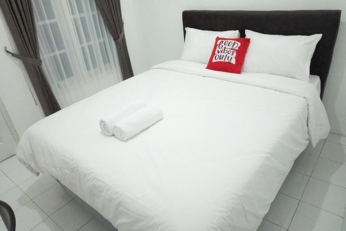 A neatly made bed with white linens and a rolled towel, a red pillow with text, and a window with curtains.