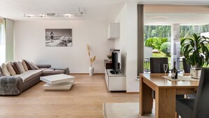 A modern living room with a sectional sofa, a flat-screen TV, a dining table with chairs, and a view of greenery outside. - Apartment "Robi" with Mountain View, Garden & Wi-Fi (Fulpmes)