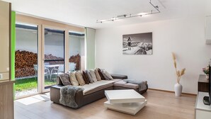 A modern living room with a sectional sofa, a coffee table, and a large window offering a view of a patio area with outdoor furniture. - Apartment "Robi" with Mountain View, Garden & Wi-Fi (Fulpmes)