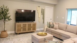 Una sala de estar con una televisión de pantalla plana sobre un mueble de madera, un sofá color beige y una planta en maceta.