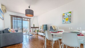 Una sala moderna con un sofá gris, sillas blancas, una mesa de comedor para cuatro comensales y un ventanal con cortinas.