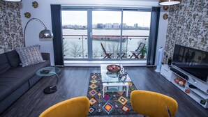 A modern living room with a sofa, a glass coffee table, a TV, and a view of a river and cityscape through large windows. - Dejayz Apartment (London)