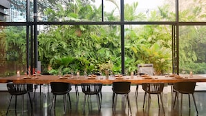 A long dining table set for six with chairs, surrounded by large windows offering a view of lush greenery.