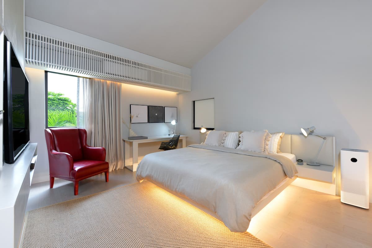 A modern hotel room with a large bed, a red chair, a desk with a computer, a TV, and a large window with curtains.