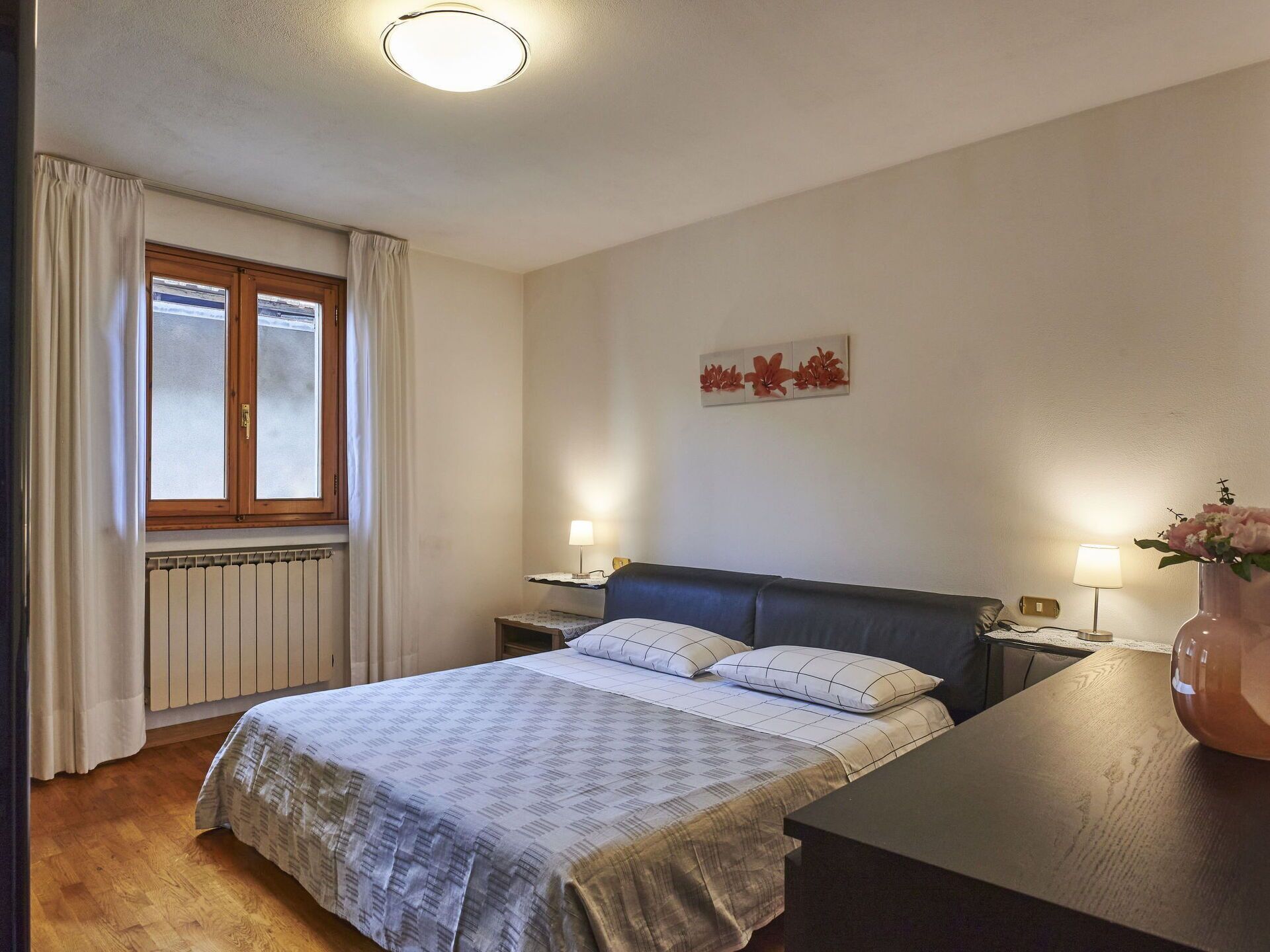 Un dormitorio con cama, mesitas de noche, una ventana con cortinas, un radiador y una obra de arte en la pared.