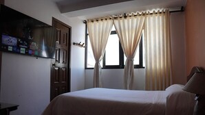 Une chambre d’hôtel comprenant un lit, un téléviseur à écran plat fixé au mur, une fenêtre avec des rideaux et une porte.