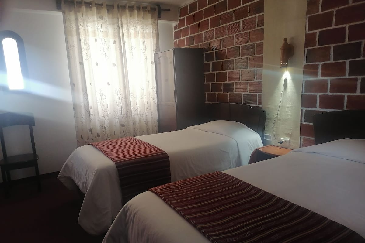 Habitación de hotel con dos camas, una pared de ladrillo, una ventana con cortinas, una silla y una mesita de noche con una lámpara.