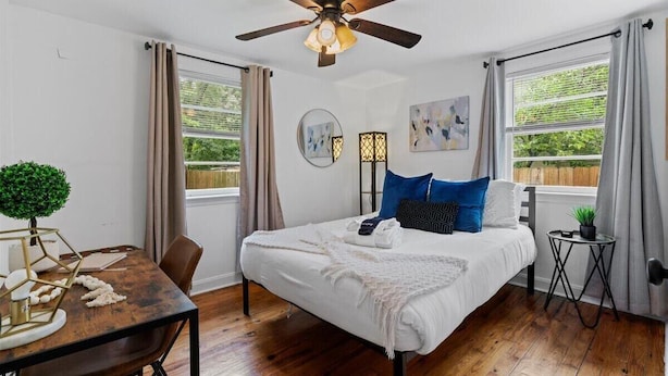 Quarto com cama, ventilador de teto, uma mesinha com uma planta e janela com cortinas.