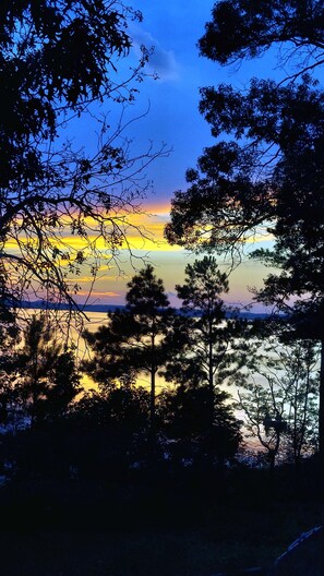 Une vue du coucher de soleil à travers les branches d’un arbre, avec une étendue d’eau calme et des collines lointaines.
