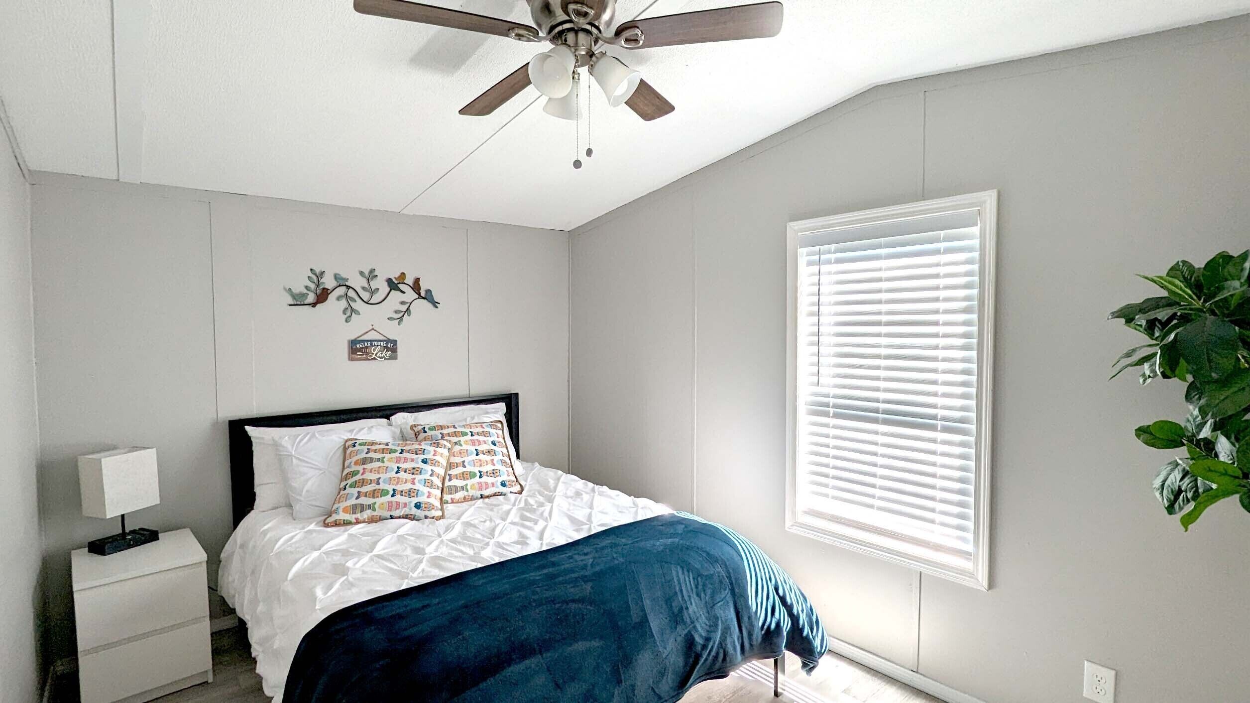 A bedroom with a bed, a nightstand, a ceiling fan, a window with blinds, and a wall-mounted decoration.