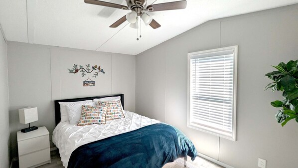 Ein Schlafzimmer mit Bett, Nachttisch, Deckenventilator, Fenster mit Jalousien und Wanddeko.