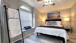 Une chambre à coucher avec un lit, deux lampes de chevet, un ventilateur de plafond, une fenêtre avec des stores et un tableau au mur.