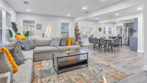 A spacious living room with a gray sofa, coffee table, and a colorful rug. There is a dining area with a table and chairs, and a kitchen in the background.