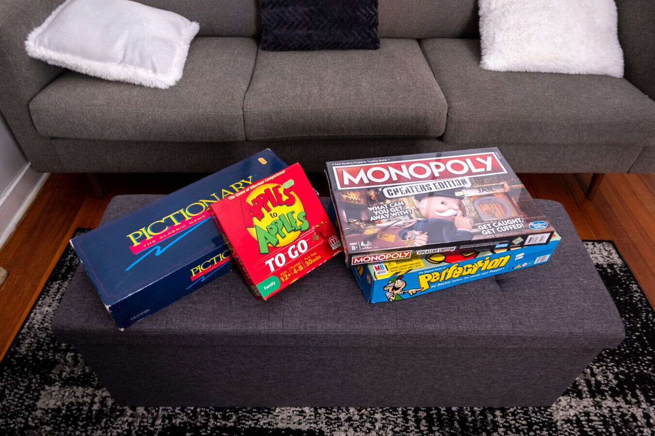 A grey sofa with white and black cushions, a black ottoman, and three board games: Pictionary, Apples to Apples, and Monopoly.