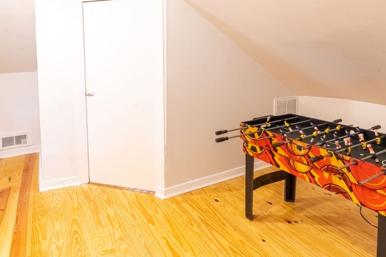 A room with a wooden floor, a white door, and a foosball table with a colorful tablecloth.