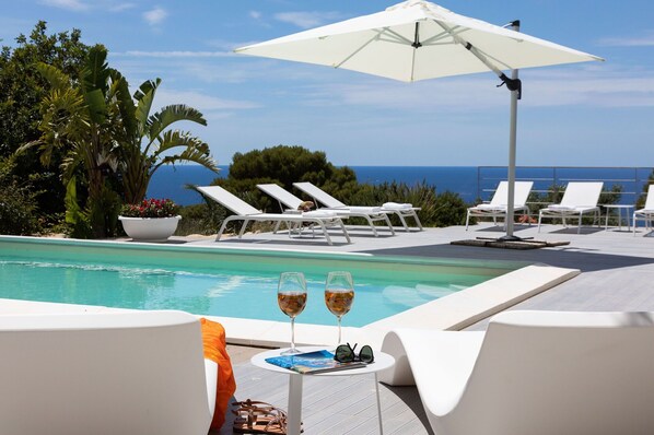 Zona de piscina con sillones de descanso, una mesa con bebidas y vista al mar.