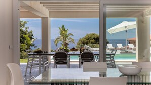 Un área de comedor al aire libre, moderna, con mesa y sillas de vidrio, con vista a una piscina y sillas de descanso.