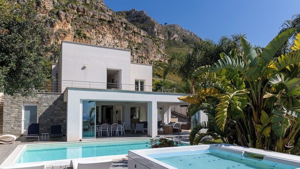 Una villa moderna con piscina, mobiliario de exterior y montañas al fondo.