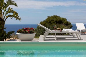 Un área junto a la piscina con sillones de descanso, una planta en maceta con flores rojas y vista al mar.
