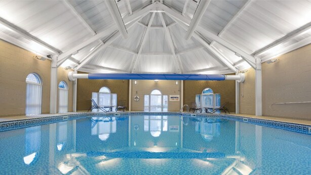 An indoor swimming pool with a tiled floor, white ceiling beams, and a blue border around the pool.