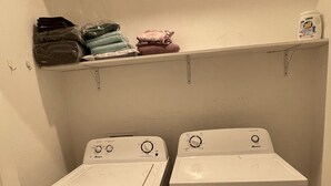 A laundry room with two front-loading washing machines and a shelf above holding folded clothes and a detergent bottle. - Loft 654B two blocks from BART (Richmond)