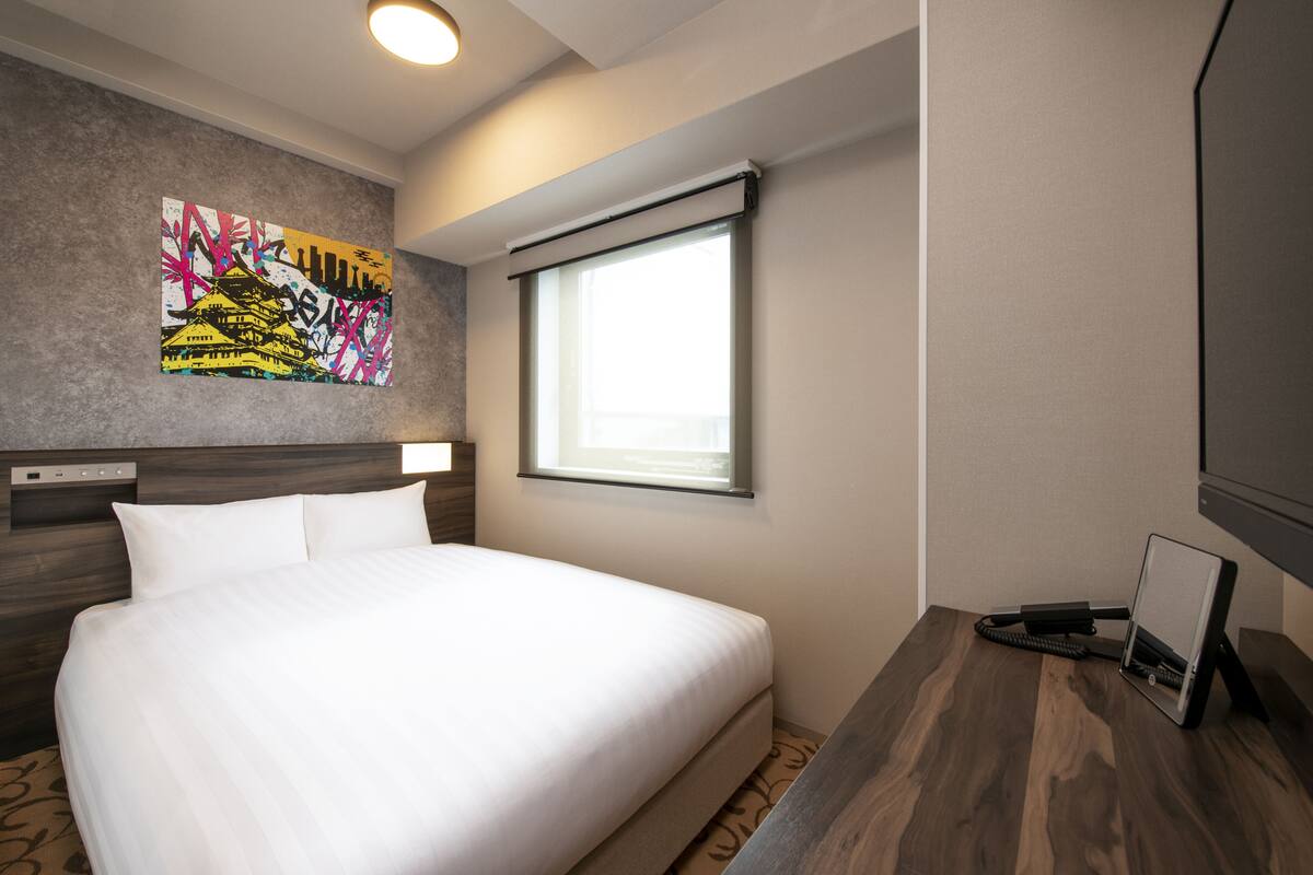 Habitació d'hotel amb un llit, una taula de treball amb un ordinador, una finestra amb persianes i una obra d'art penjada a la paret.