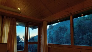 Una habitación de cabaña de madera con una cama, un ventanal con vistas a un bosque y una lámpara en el techo.
