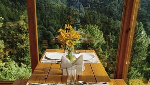 Una mesa de madera dispuesta para dos personas, con un jarrón de girasoles, frente a un paisaje montañoso.