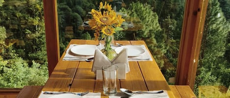 A wooden table set for two with a vase of sunflowers, set against a backdrop of a mountainous landscape.