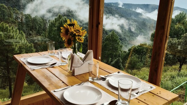 Mesa de madera con platos, cubiertos y un vaso de agua, con vista a un paisaje montañoso con vegetación exuberante y neblina.