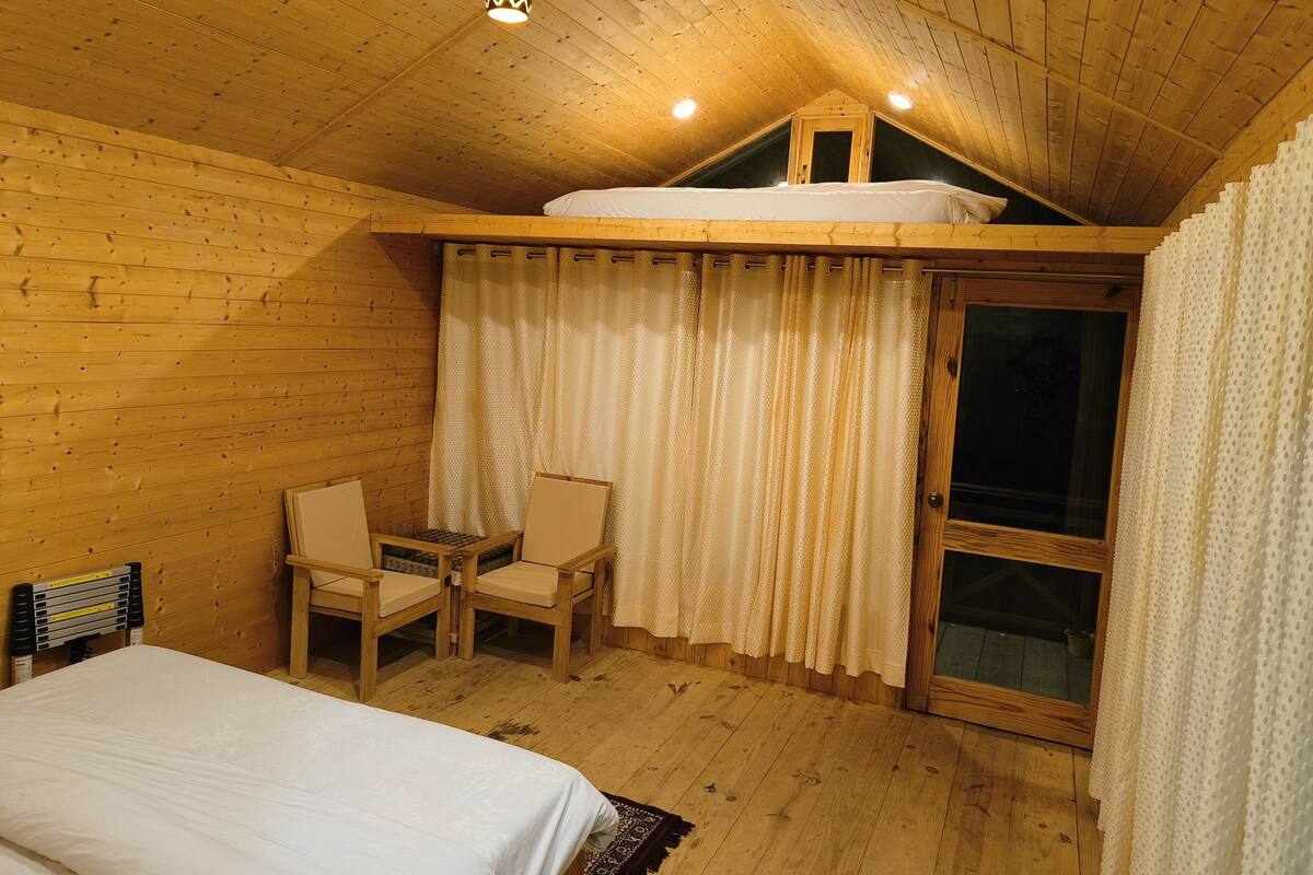 A cozy attic bedroom with a wooden ceiling, a bed, a desk, two chairs, and a window with curtains.