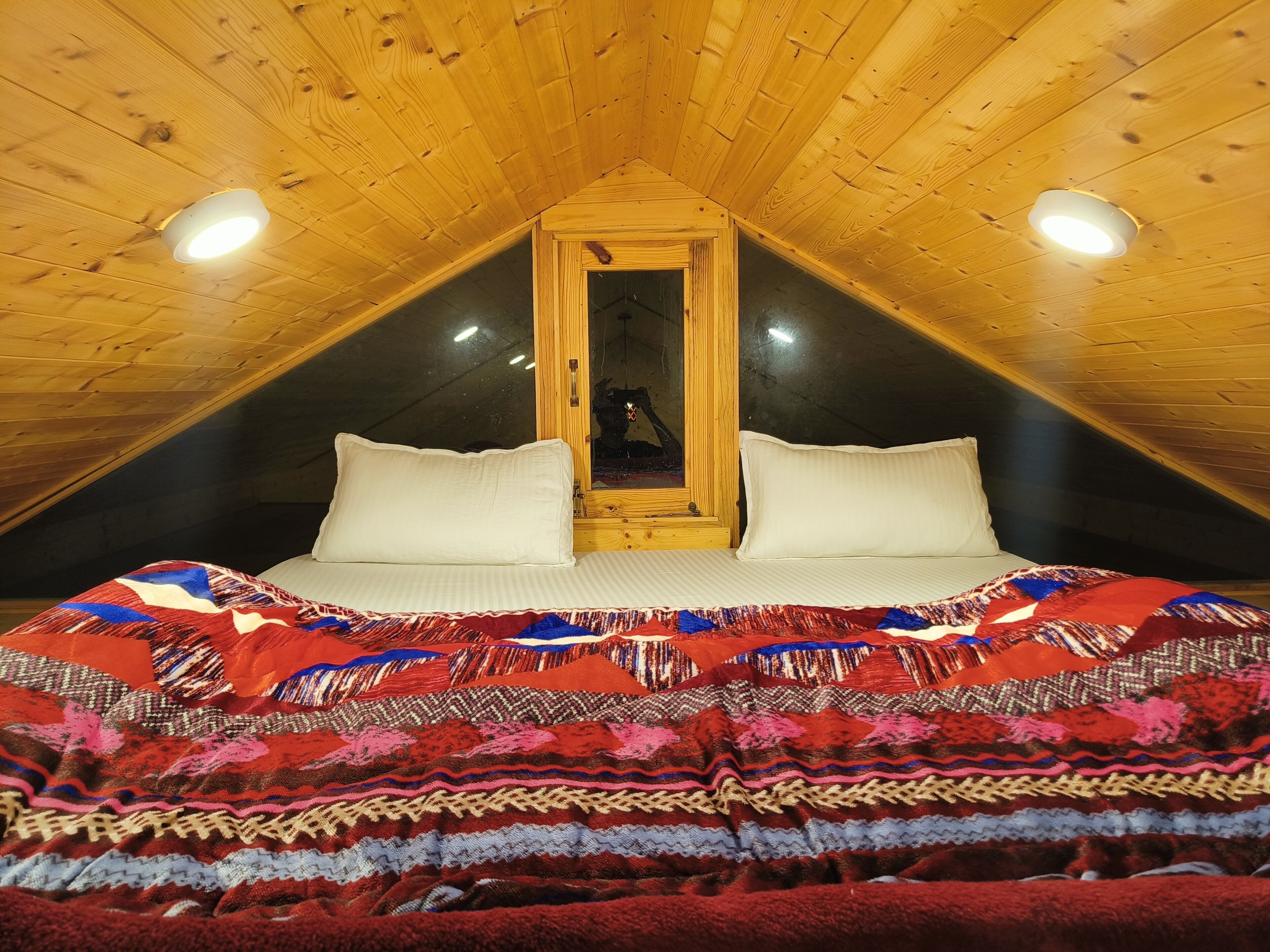 Een knus slaapkamer onder een schuin dak, met een groot bed en een kleurrijk dekbed, en een houten hoofdbord.