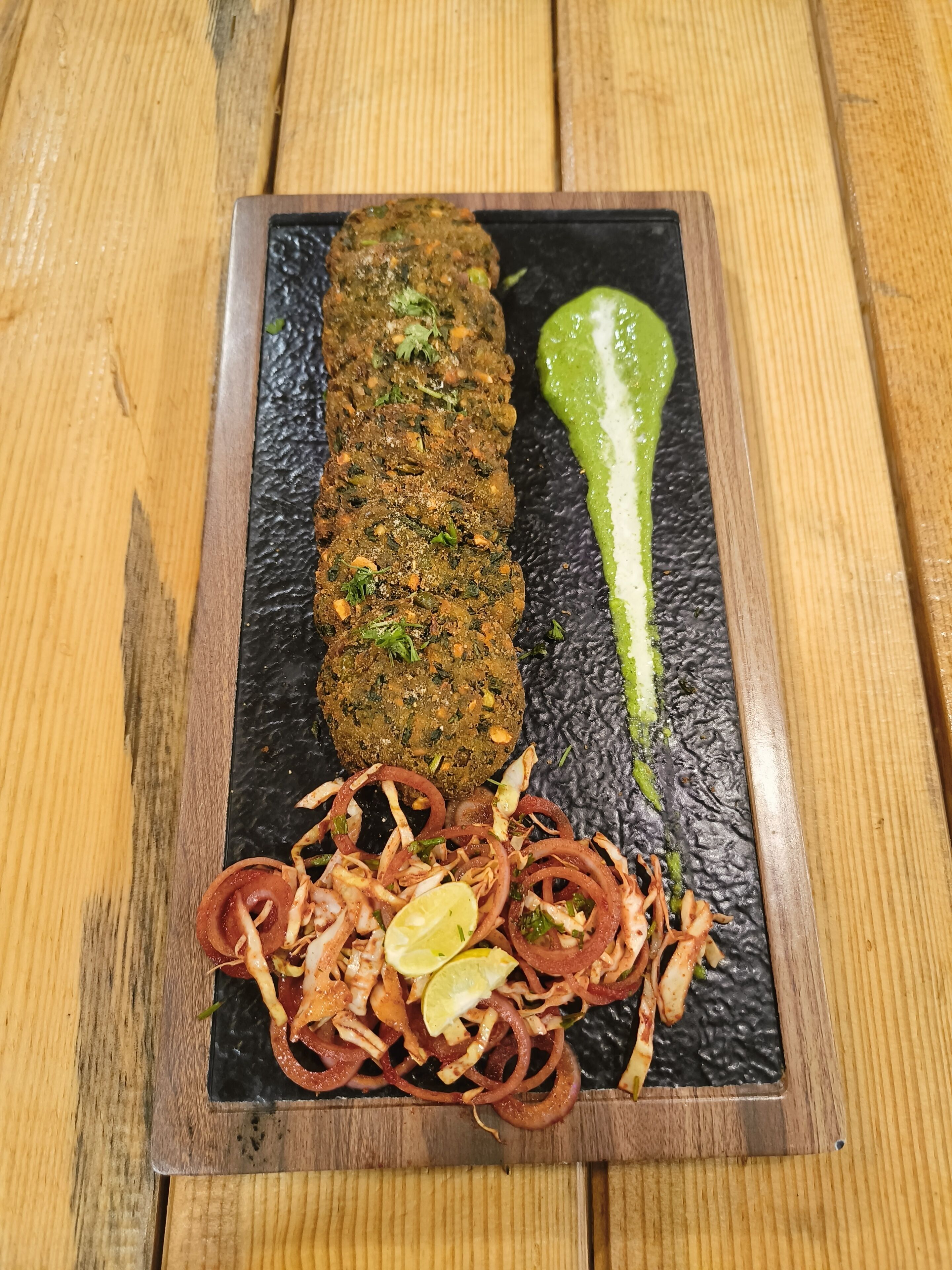 A rectangular black slate serving tray with a wooden border, containing a meat dish, a green sauce, and a side of thinly sliced onions.