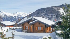 Una cabana de fusta amb un teulat cobert de neu, envoltada de terreny nevós i muntanyes al fons.