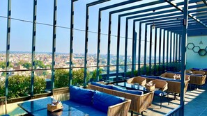 A rooftop lounge with wicker furniture and a glass table, overlooking a cityscape. - Hotel Ganga Residency (Ganganagar)